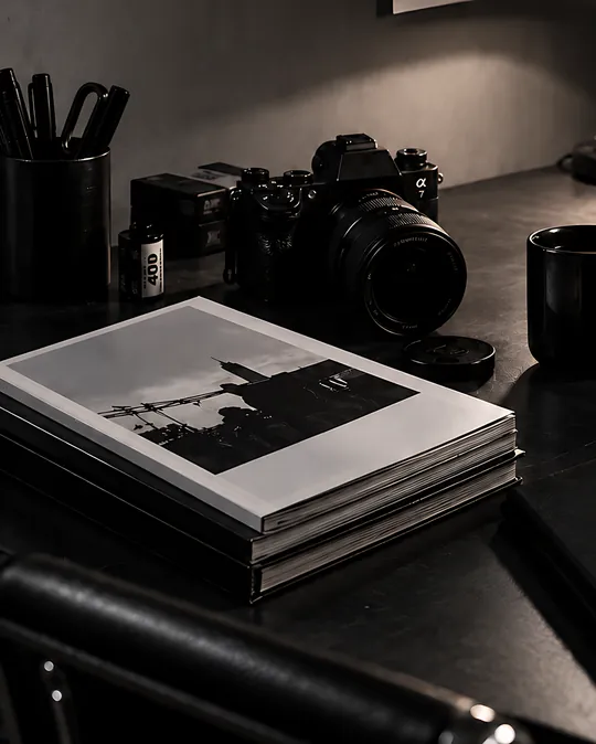 A camera, a stack of photo books and a cup of coffee on a dark desk under a lamp.