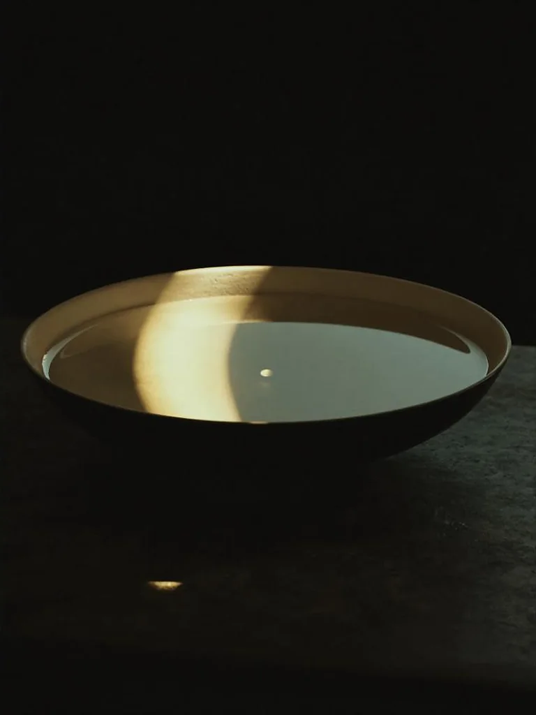 A shallow ceramic bowl of perfectly still water on dark stone, crossed by a single narrow shaft of warm gold light, the surface completely unbroken.