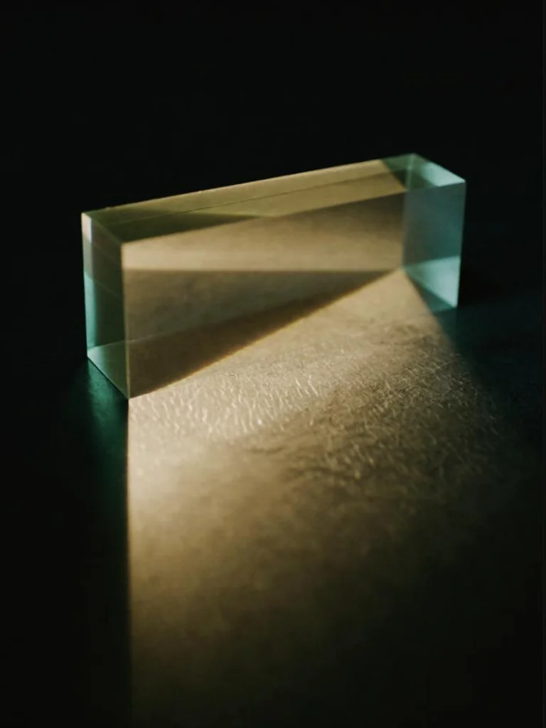 A solid block of optical glass on a dark surface, gathering scattered warm light and laying it down as one clean resolved beam.